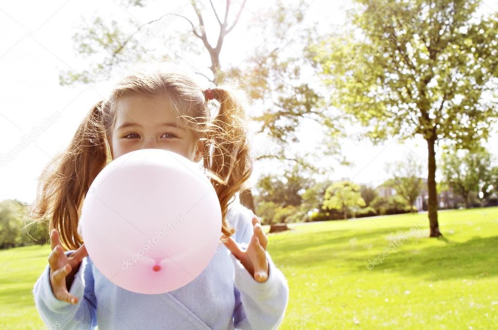 Young girl blowing a pink balloon in the park on a sunny day.