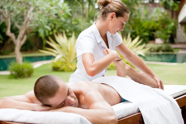 Young masseuse massaging and stretching the body of an attractive man in a tropical hotel garden near a swimming pool. — Stock Photo, Image