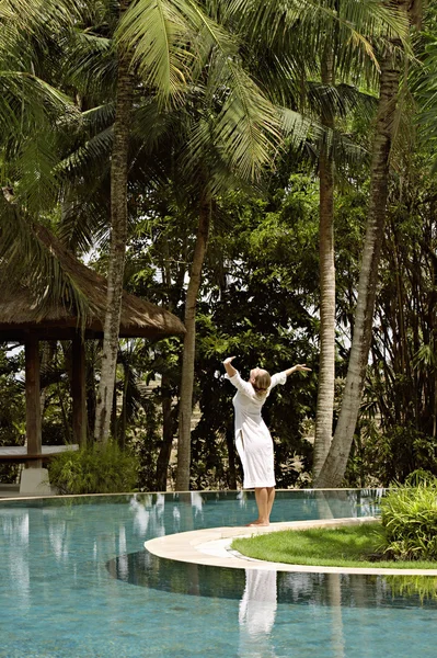 Young woman standing at the edge of a swimming pool surrounded by tall palm trees — Stock Photo, Image