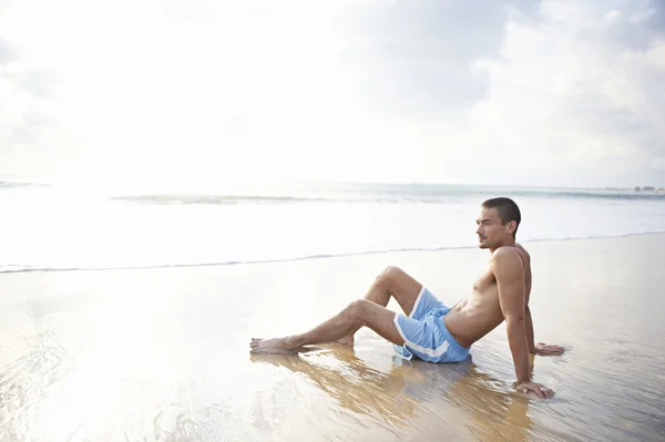 Young attractive man sitting down on the wide shore of a golden sand beach Stock Photo