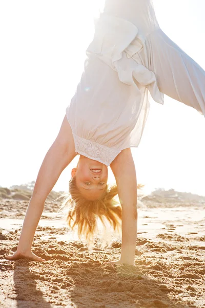 Jong meisje doen cartwheels op een gouden zand strand met de stralen van de zon filteren door middel van haar lichaam tijdens zonsondergang. — Stockfoto