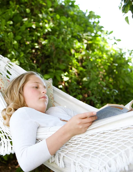 Ung tonårsflicka som läser en bok medan fastställande av i en hängmatta i en trädgård. — Stockfoto