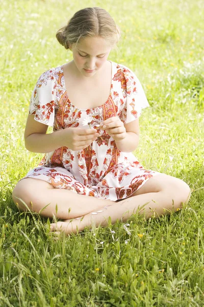 Girl pulling petals off a daisy flower while sitting on a green field, playing love me, love me not. — Stock Photo, Image