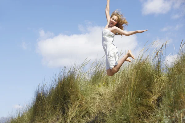 Girl jumping off a long grass hill on the beach, with a blue sky in the background. Stock Image