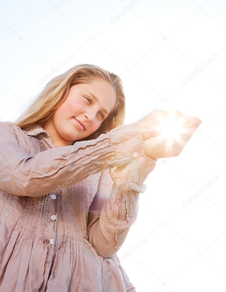 Young blond girl holding the sun in her hands, with sunrays filtering through, against a blue sky.