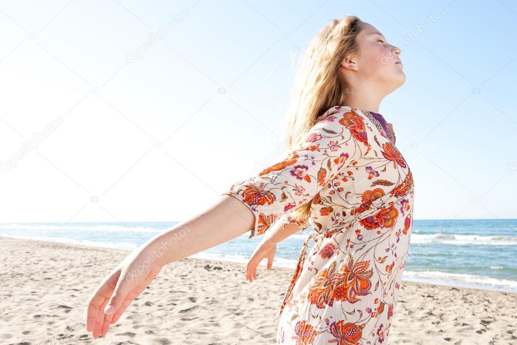 Young girl breathing fresh air while enjoying the sun on a golden sand beach with a blue sky and the sea horizon in the background.