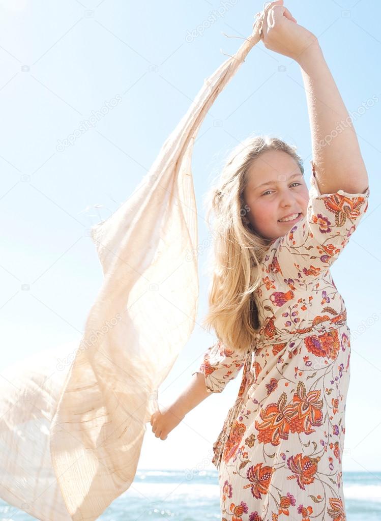 Young teenage girl holding a sarong in the air blowing in the wind against a blue sky, smiling.