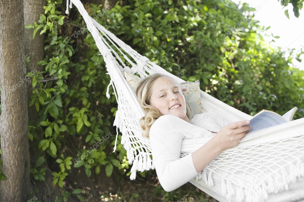 Girl laying down on a hammock in the garden, reading a book and smiling.