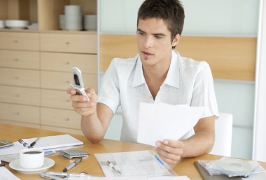 ' ın evinin mutfağında maliyesi üzerinde çalışırken, cep telefonu kullanan adam.