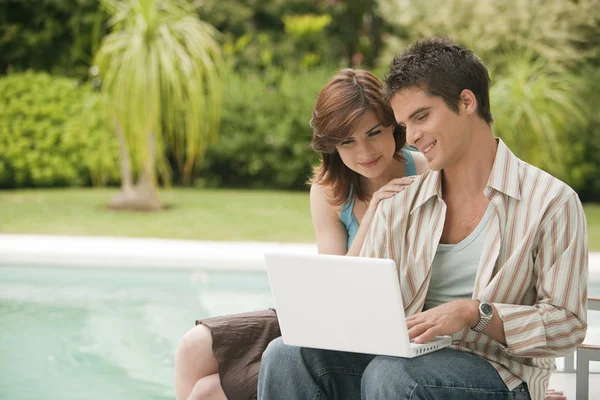 Pareja usando un ordenador portátil en casa junto a la piscina, exterior . — Foto de Stock