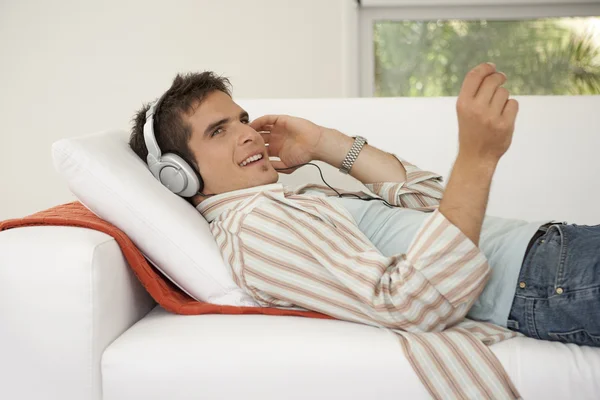 Vista lateral del hombre acostado en un sofá blanco escuchando música en casa . — Foto de Stock