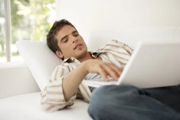 Mann benutzt Laptop, während er sich zu Hause auf ein weißes Sofa legt. — Stockfoto