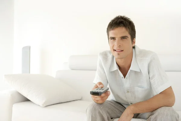 Hombre joven usando un control remoto de televisión mientras está sentado en un sofá de cuero blanco en casa . — Foto de Stock