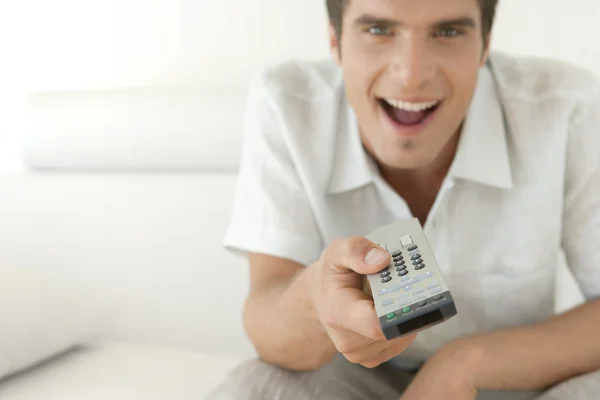 Ung man med en TV-kontrollen remote medan du sitter på en vit skinnsoffa hemma. — Stockfoto