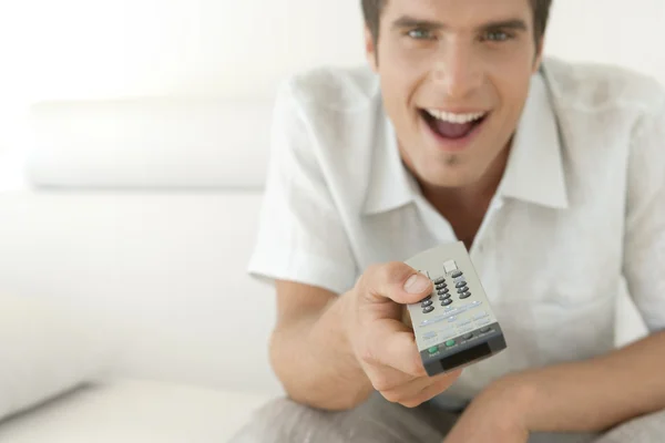 Hombre joven usando un control remoto de televisión mientras está sentado en un sofá de cuero blanco en casa . —  Fotos de Stock