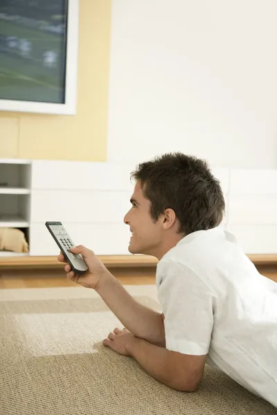 Jovem profissional usando um controle remoto de TV enquanto assiste a uma tv de tela plana em casa . — Fotografia de Stock