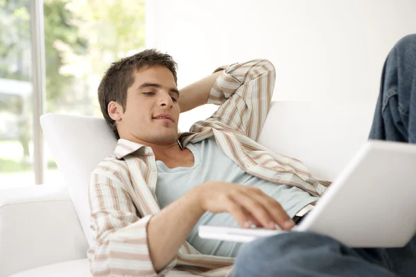 Hombre usando una computadora portátil mientras está acostado en un sofá blanco en casa . —  Fotos de Stock