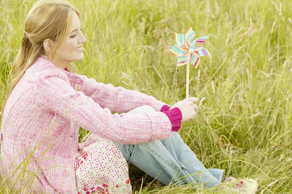 Jeune femme tenant un moulin à vent coloré tout en s'asseyant sur une longue herbe verte — Photo