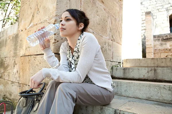 young businesswoman sitting on a park steps drinking water from a plastic blue bottle.