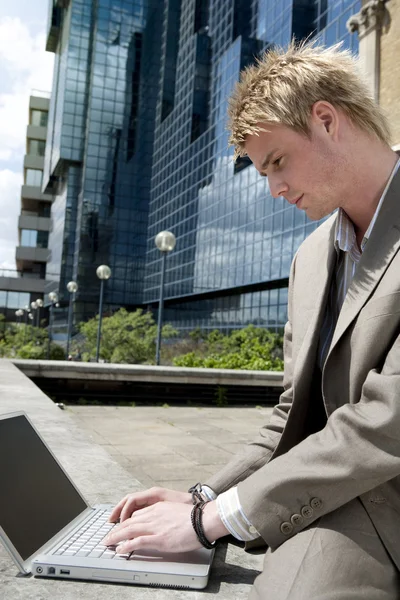 Jovem empresário digitando em um computador portátil enquanto está sentado perto do prédio de escritórios moderno . — Fotografia de Stock