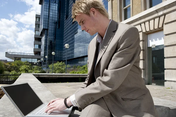 Jovem empresário digitando em um computador portátil enquanto está sentado perto do prédio de escritórios moderno — Fotografia de Stock