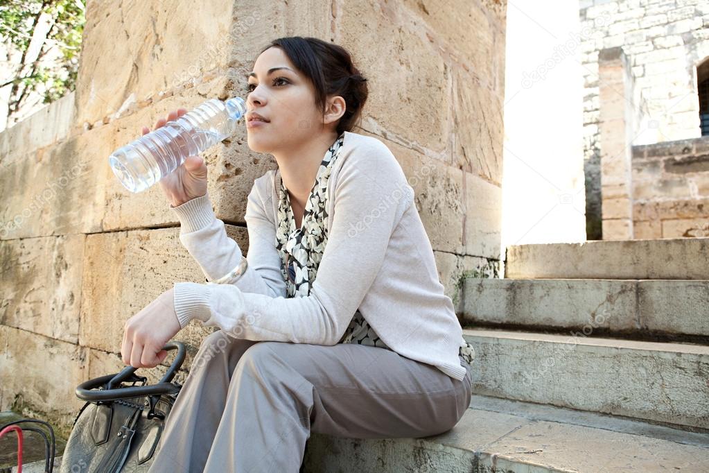 young businesswoman sitting on a park steps drinking water from a plastic blue bottle.