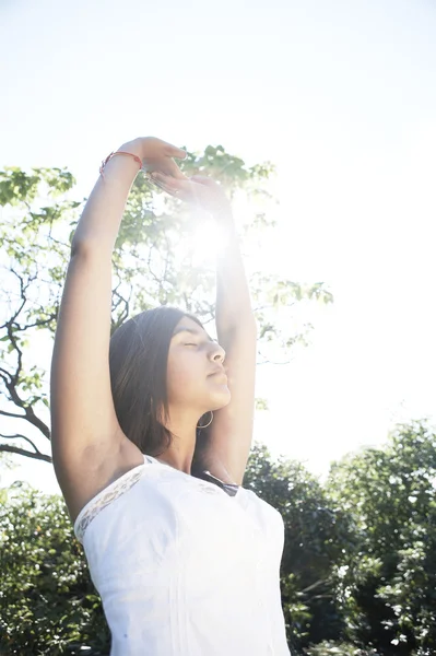 Young indian woman stretching and doing yoga in the park with the sun filtering through her arms. — Stock Photo, Image