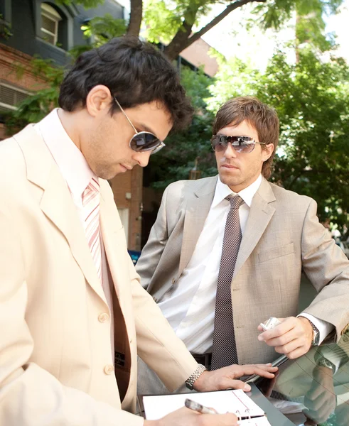 Two businessmen working while leaning on a car in a tree lined street in the city. — Stock Photo, Image