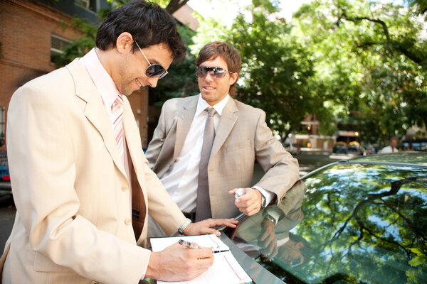 Two businessmen working while leaning on a luxury car in a tree lined street in the city.