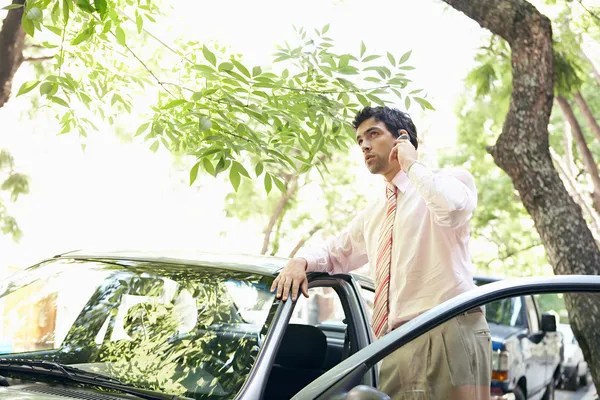 Empresário atraente apoiando-se no topo de um carro enquanto faz um telefonema em uma rua frondosa . — Fotografia de Stock