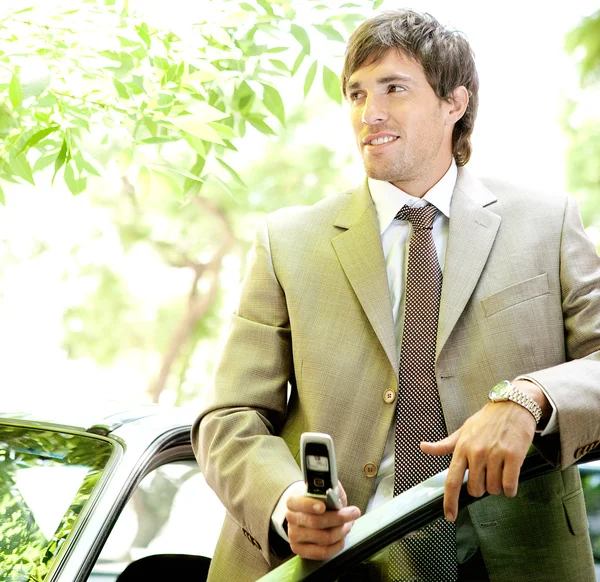 Joven hombre de negocios sosteniendo un teléfono celular mientras se apoya en la puerta de su coche — Foto de Stock