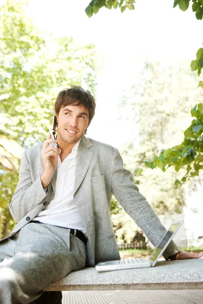 Jovem empresário atraente usando um telefone celular e um computador portátil — Fotografia de Stock