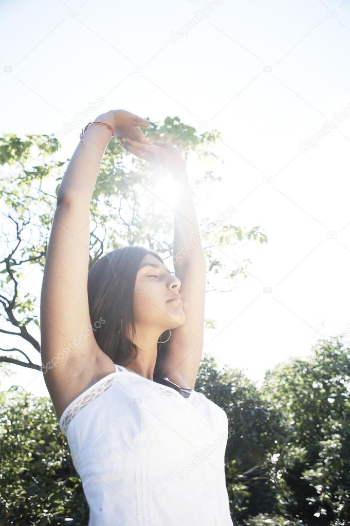 Young indian woman stretching and doing yoga in the park with the sun filtering through her arms.