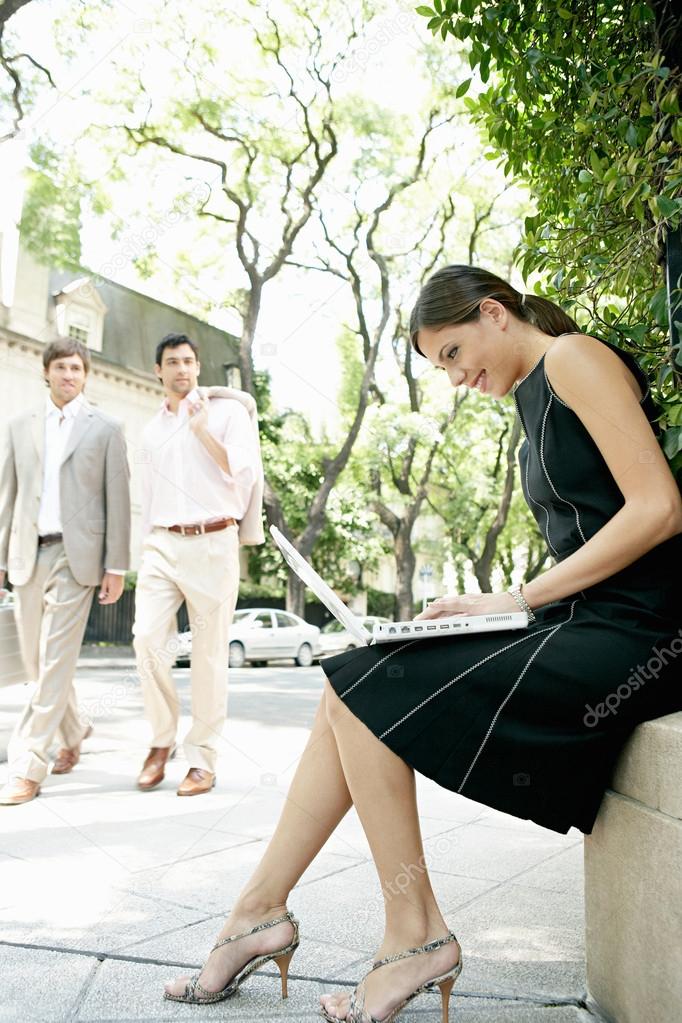 Two businessmen walking in the street and looking at a sexy businesswoman sitting down using her laptop computer.