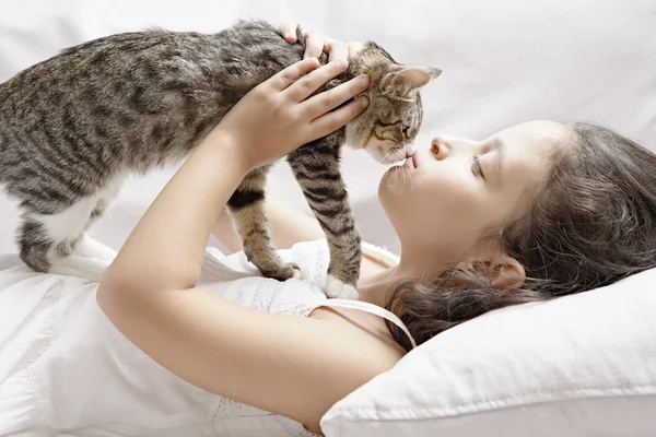 Young girl kissing kitten while laying down on a white sofa at home. — Stock Photo, Image