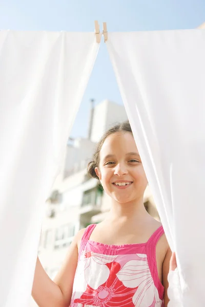 Menina sorrindo para a câmera e segurando lençóis brancos enquanto eles secam pendurados em um terraço . — Fotografia de Stock