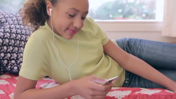 Молодая девушка-подросток пролистывает свой сенсорный экран смартфона и поворачивается, чтобы улыбнуться в камеру, лежа на диване дома . — стоковое видео