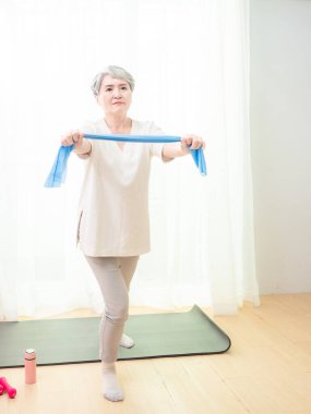 Kıdemli Asyalı kadın evde direniş bandıyla egzersiz yapıyor..