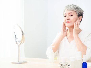 Mutlu 50 'ler orta yaşlı Asyalı kadın aynadaki yansımaya bakarak tenine dokunuyor. Gülümseyen yaşlı kadın şımartıyor, sağlıklı nemlendirilmiş cilt bakımı, yaşlanan güzellik, cilt bakımı kozmetik konsepti.