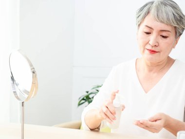 Mutlu 50 'ler orta yaşlı Asyalı kadın aynadaki yansımaya bakarak tenine dokunuyor. Gülümseyen yaşlı kadın şımartıyor, sağlıklı nemlendirilmiş cilt bakımı, yaşlanan güzellik, cilt bakımı kozmetik konsepti.