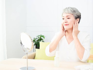 Mutlu 50 'ler orta yaşlı Asyalı kadın aynadaki yansımaya bakarak tenine dokunuyor. Gülümseyen yaşlı kadın şımartıyor, sağlıklı nemlendirilmiş cilt bakımı, yaşlanan güzellik, cilt bakımı kozmetik konsepti.