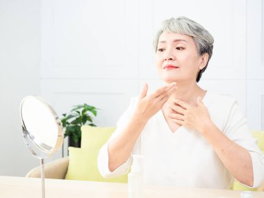 Mutlu 50 'ler orta yaşlı Asyalı kadın aynadaki yansımaya bakarak tenine dokunuyor. Gülümseyen yaşlı kadın şımartıyor, sağlıklı nemlendirilmiş cilt bakımı, yaşlanan güzellik, cilt bakımı kozmetik konsepti.