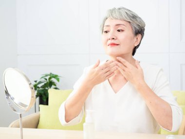 Mutlu 50 'ler orta yaşlı Asyalı kadın aynadaki yansımaya bakarak tenine dokunuyor. Gülümseyen yaşlı kadın şımartıyor, sağlıklı nemlendirilmiş cilt bakımı, yaşlanan güzellik, cilt bakımı kozmetik konsepti.