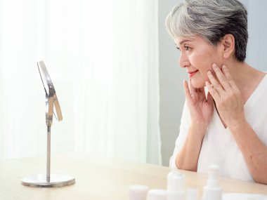 Mutlu 50 'ler orta yaşlı Asyalı kadın aynadaki yansımaya bakarak tenine dokunuyor. Gülümseyen yaşlı kadın şımartıyor, sağlıklı nemlendirilmiş cilt bakımı, yaşlanan güzellik, cilt bakımı kozmetik konsepti.