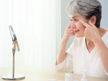 Mutlu 50 'ler orta yaşlı Asyalı kadın aynadaki yansımaya bakarak tenine dokunuyor. Gülümseyen yaşlı kadın şımartıyor, sağlıklı nemlendirilmiş cilt bakımı, yaşlanan güzellik, cilt bakımı kozmetik konsepti.
