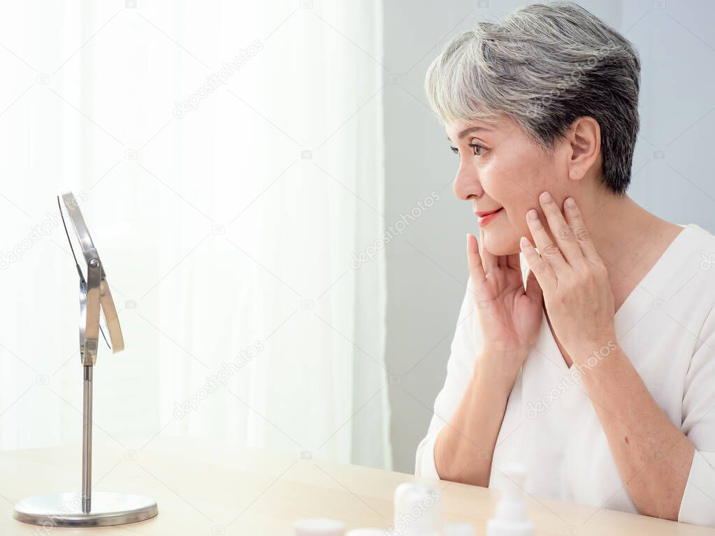 Happy 50s middle aged asian woman touching face skin looking in mirror reflection. Smiling mature old lady pampering, healthy moisturized skin care, aging beauty, skincare treatment cosmetics concept.