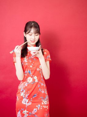 Geleneksel Cheongsam Qipao elbisesi giyen güzel Asyalı kadın yemek çubukları ve kase ile yemek yiyor, kırmızı arka planda izole edilmiş..