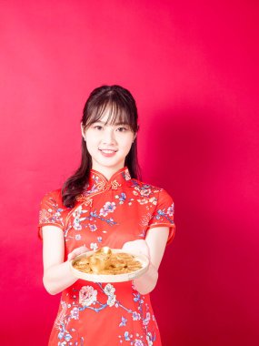 Geleneksel Cheongsam Qipao elbisesi giyen mutlu Asyalı genç kadın Çin 'in yeni yılını kırmızı arka planda kutlamak için altın külçesini gösteriyor..