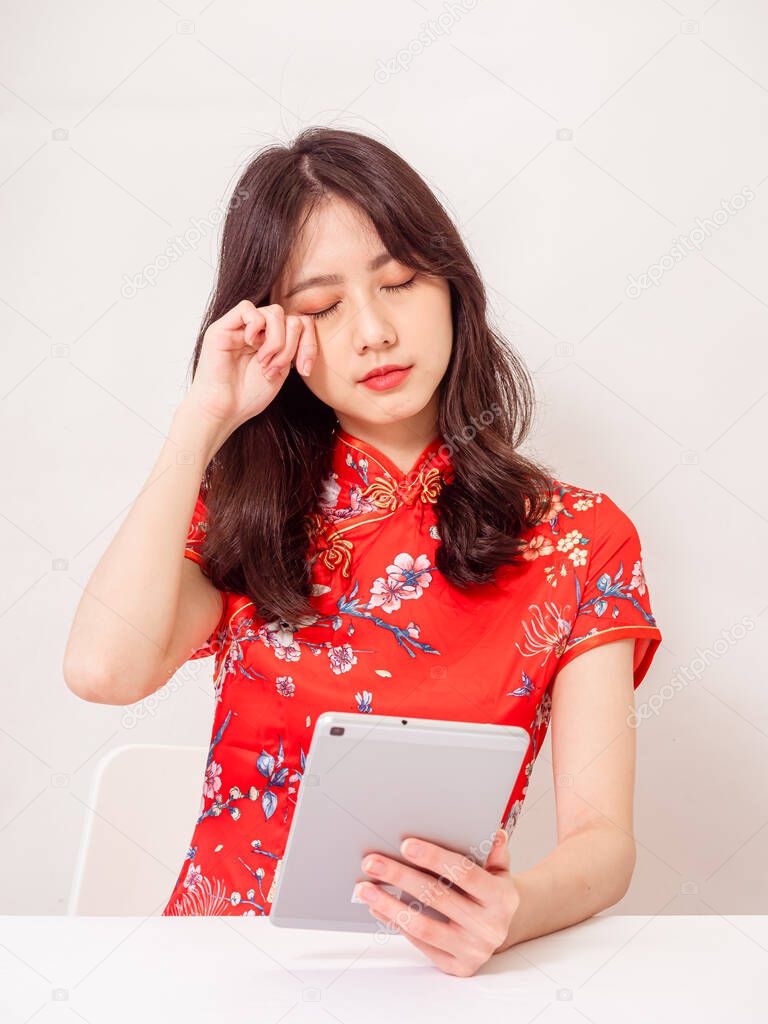 Young asian female wearing traditional cheongsam qipao dress feeling tired and upset after using tablet and rubbing eyes uncomfortably isolated on white background.