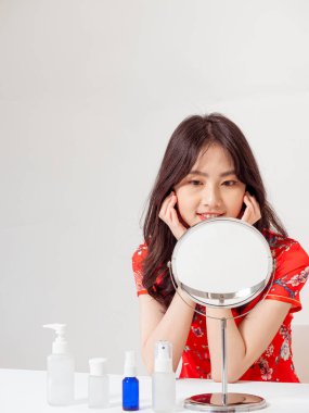 Geleneksel Cheongsam Qipao elbisesi içindeki Asyalı genç bir kadının portresi aynaya bakar ve beyaz arka planda izole edilmiş cilt bakımı yapar..